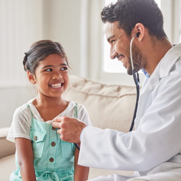 Young handsome smiling doctor using a stethoscope to listen to a sick little girls heartbeat during a house call checkup at home